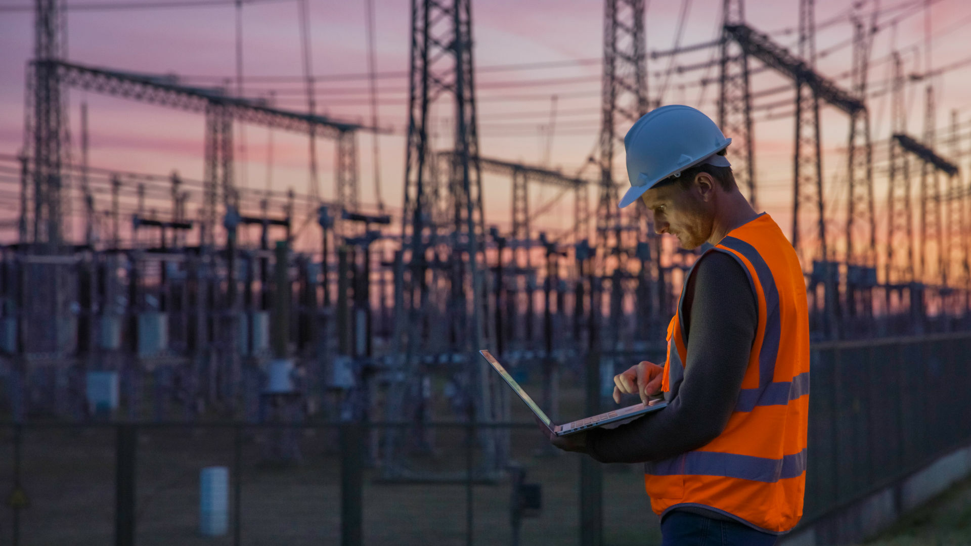 Techniker mit Laptop vor Strommasten im Abendlicht