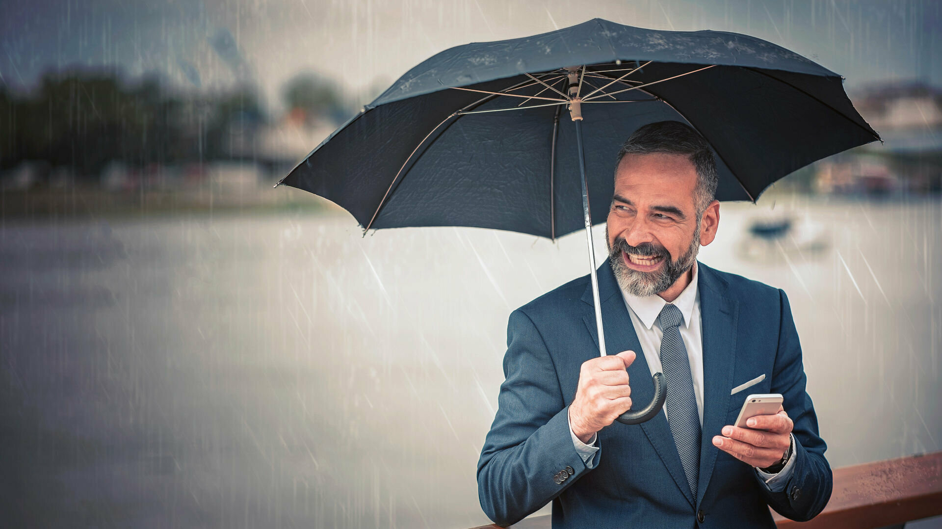 Mann steht unter einem Regenschirm und lacht und hat ein Smartphone in der Hand